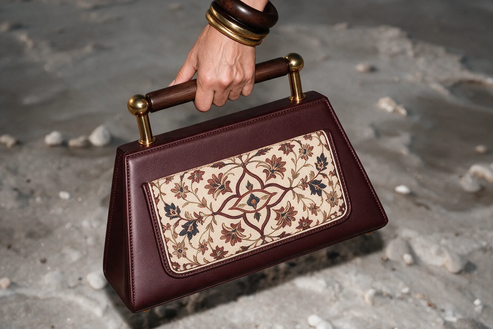 A hand with wooden and gold bangles carrying the burgundy bag with a cream floral textile panel over wet salt